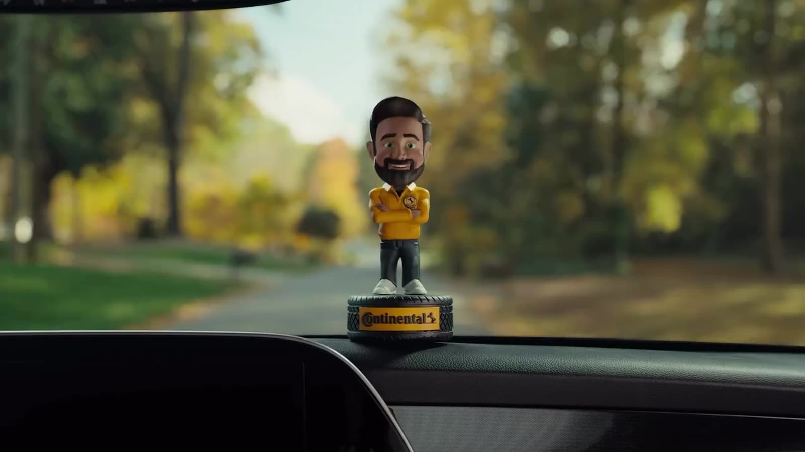 CG bobblehead on a car dashboard, autumn trees behind